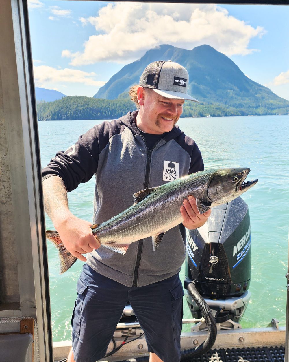 Man holding salmon on a boat with a mountain background in BC. He loves fishing charters. Book now.