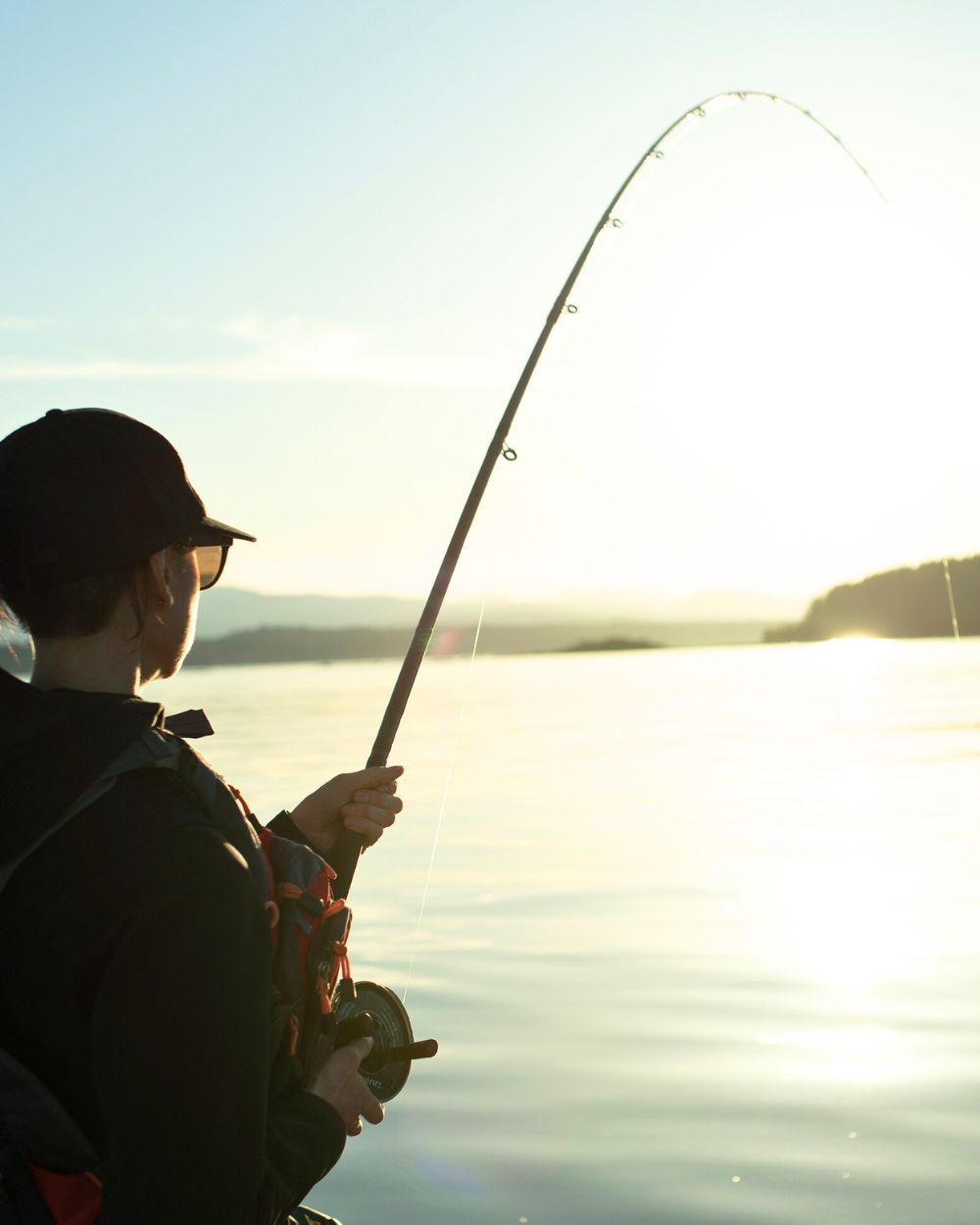 Relaxed fisherwoman with a line cast out into a BC lake with a beautiful sunset that is shimmering off of the surface of the lake in the background. She loves fishing charters. Book now.